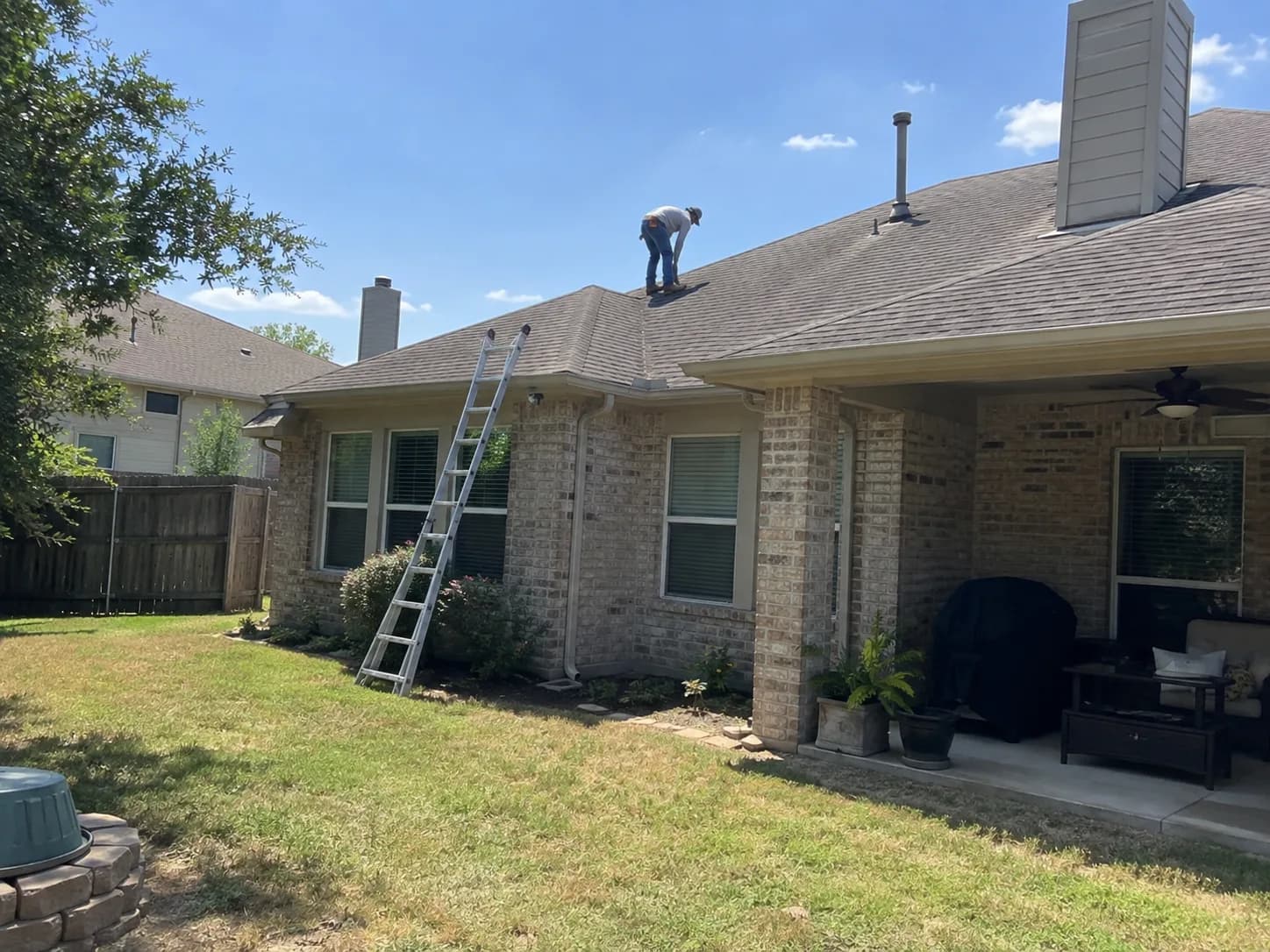 A person checking a shingle roof above a brick home's backyard