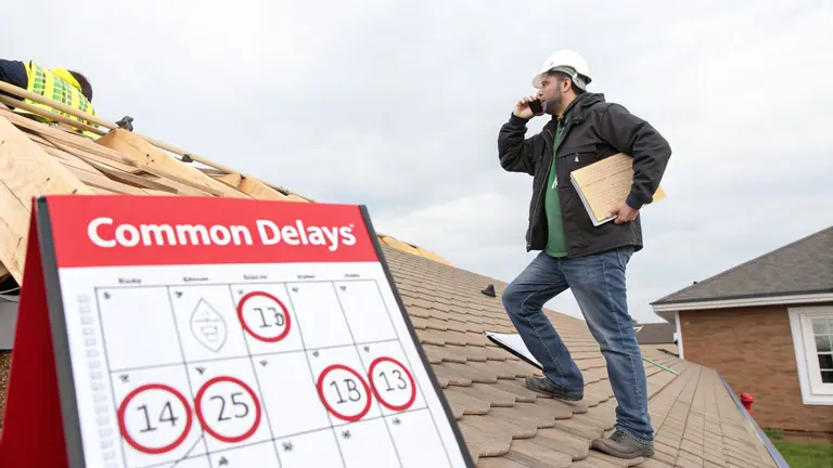 A construction manager on a residential roof talks on the phone, reviewing a 'Common Delays' sign.